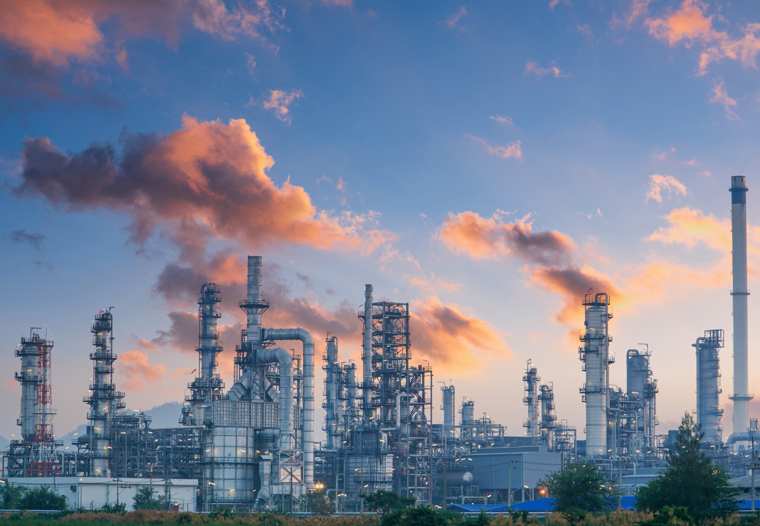 Große Chemieanlage mit zahlreichen Türmen, Rohrleitungen und Schornsteinen vor einem Himmel mit rosa-orangen Wolken während des Sonnenuntergangs.