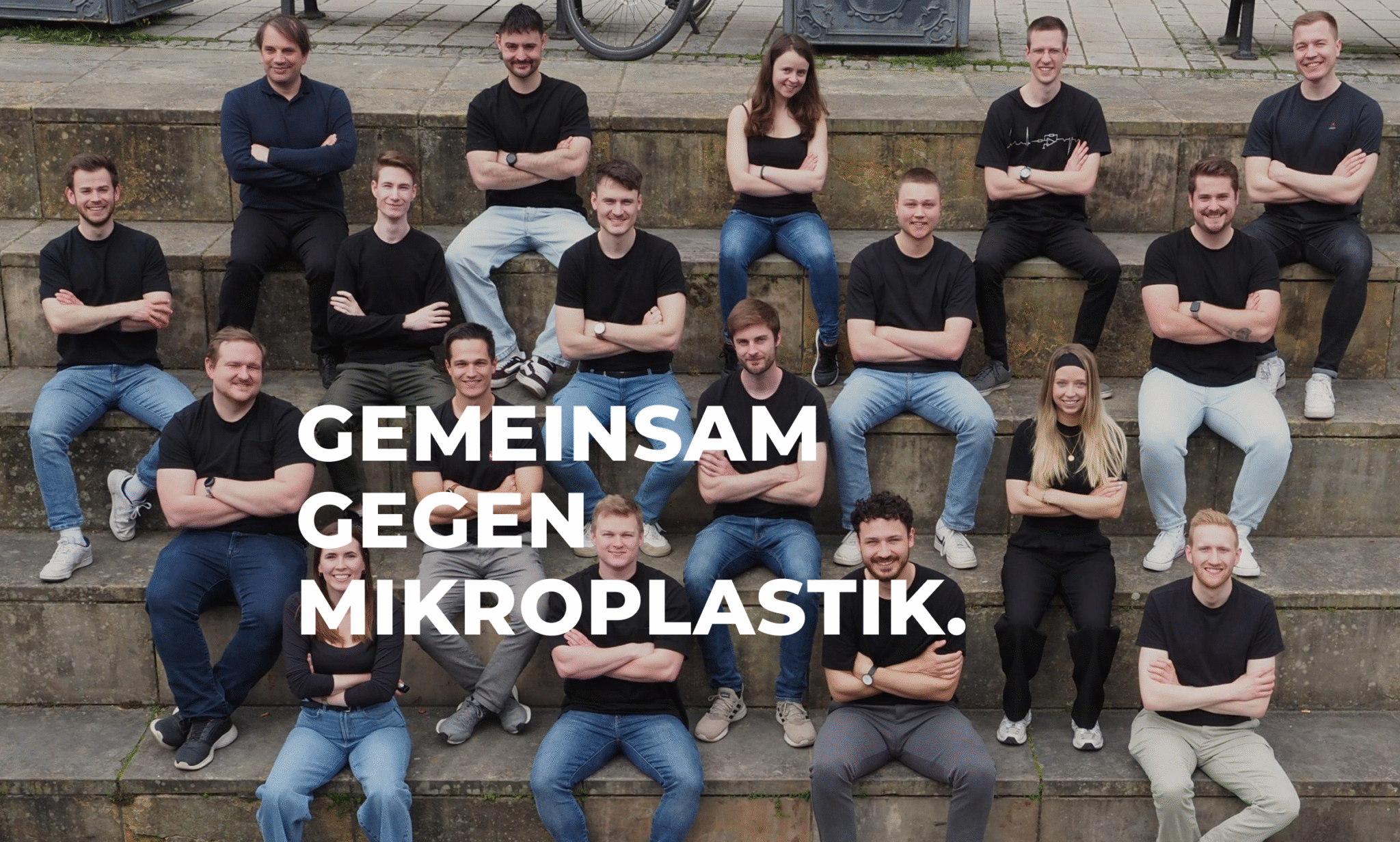 Alle Mitarbeitenden sitzen auf einer Steintreppe mit verschränkten Armen und schauen in die Kamera, die Über ihnen ist. Darüber liegt der Schriftzug: Gemeinsam gegen Mikroplastik.